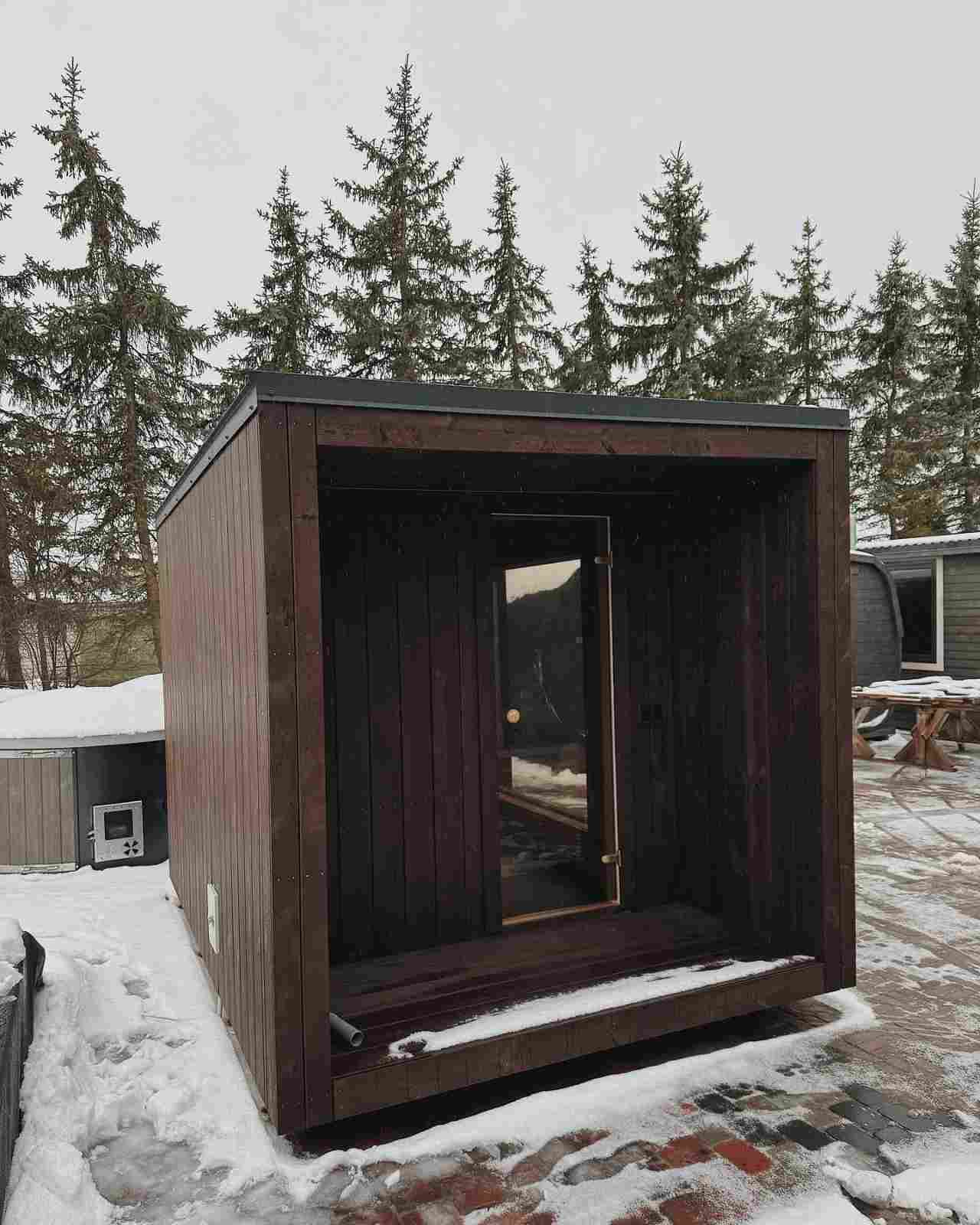 Entrance view of a minimalist dark wood cube sauna with glass door and covered porch area.