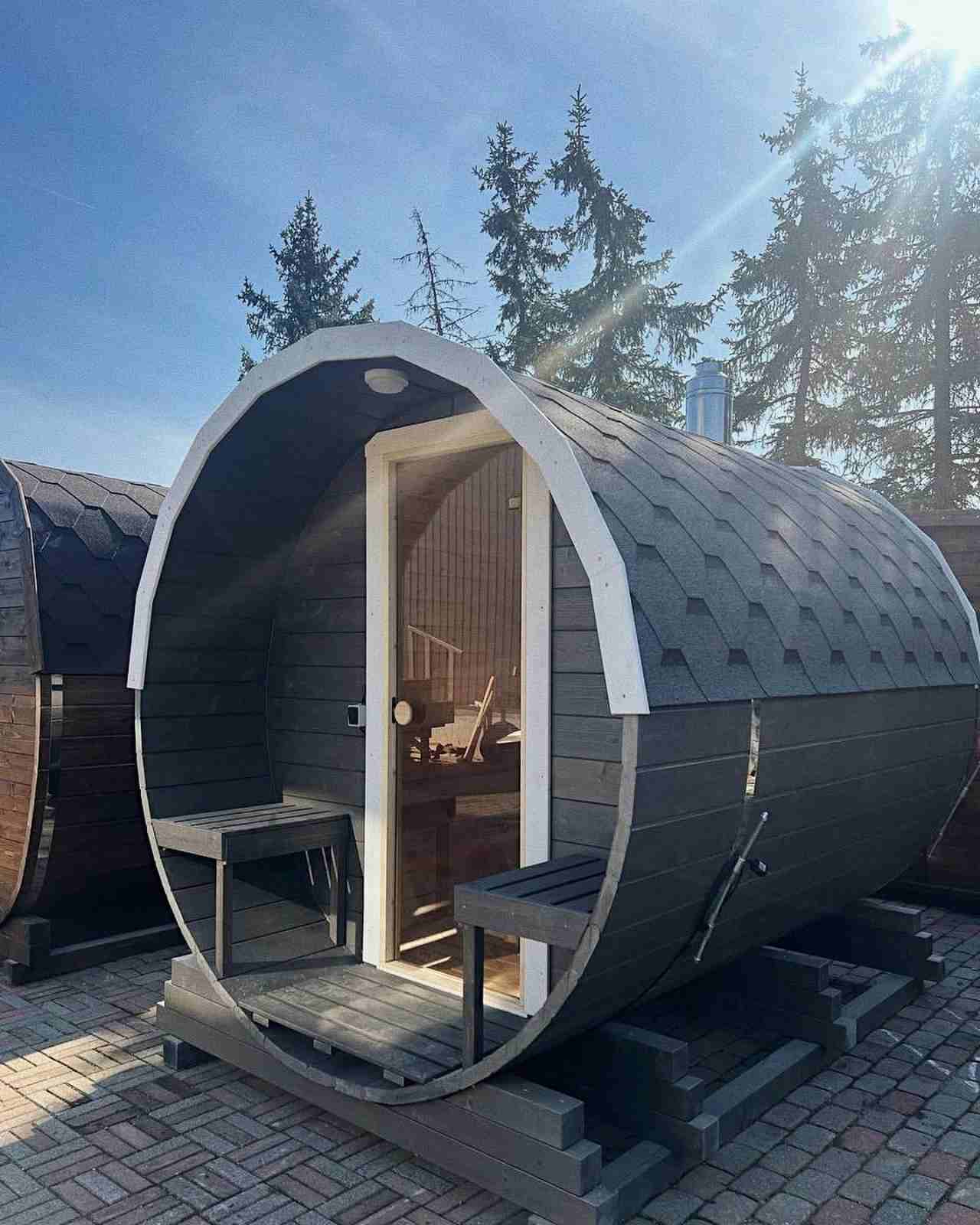 Outdoor barrel sauna with grey shingles and a small front porch area under a clear blue sky.