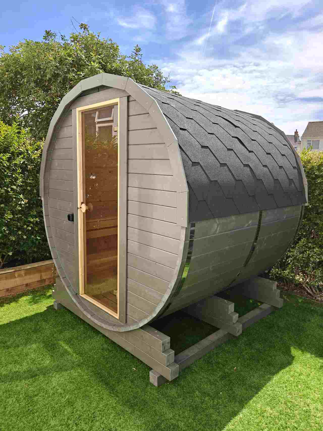 Full side view of a standalone grey barrel wooden sauna with a black shingled roof, full-length glass door, and heavy-duty timber base frame on a green grass lawn.
