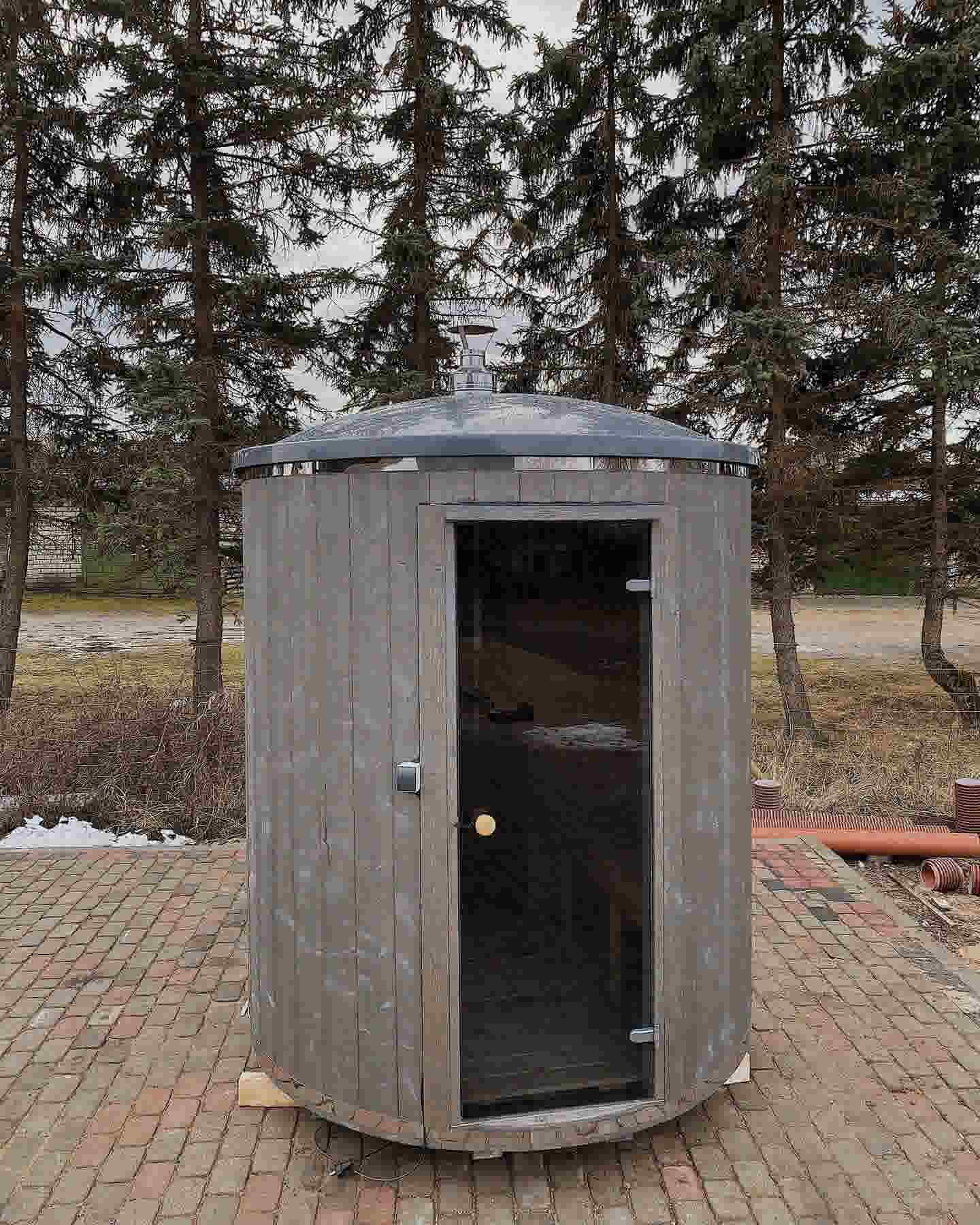 Exterior of The Cupa vertical barrel sauna in weathered grey with a dome roof and stainless steel chimney.