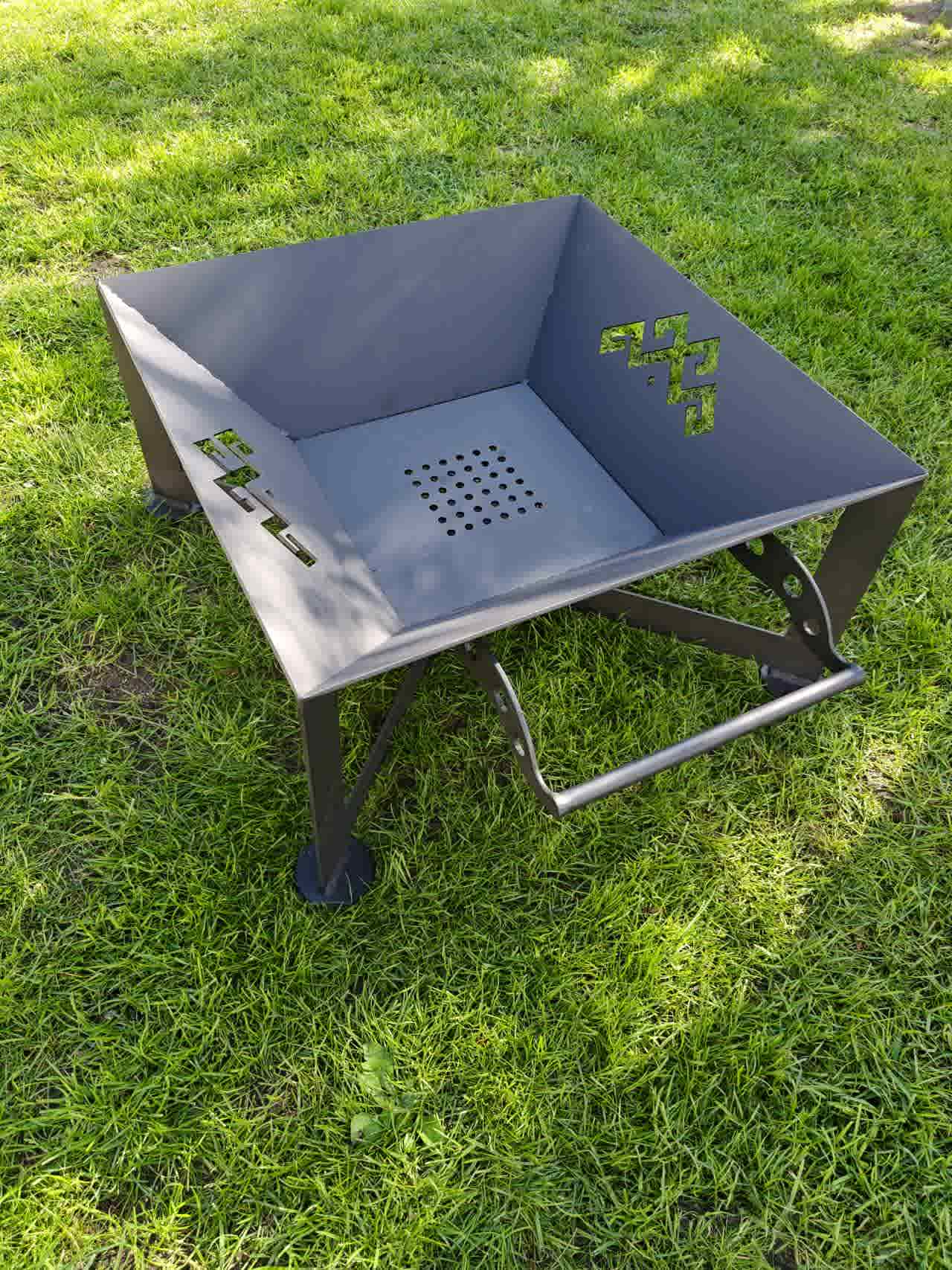 Modern square steel fire pit, The Foco, showing the architectural cross-leg base and decorative side ventilation patterns.